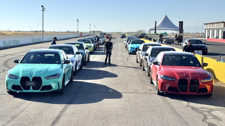 A head-on photo of all the cars lined up in the pits with some people standing around them
