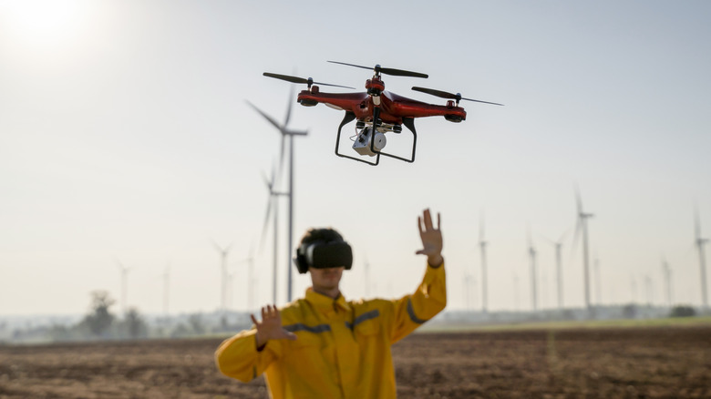 A man in a virtual reality headset and yellow jacket operates a red drone in a field of wind turbines