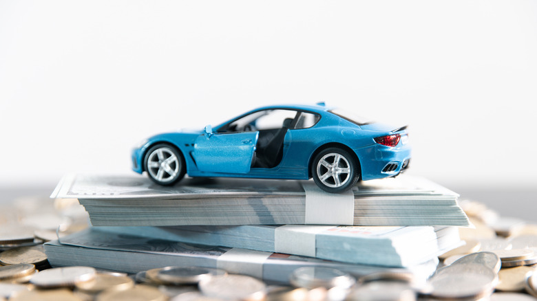 A blue Ford Mustang model car sits atop a stack of money and coins.