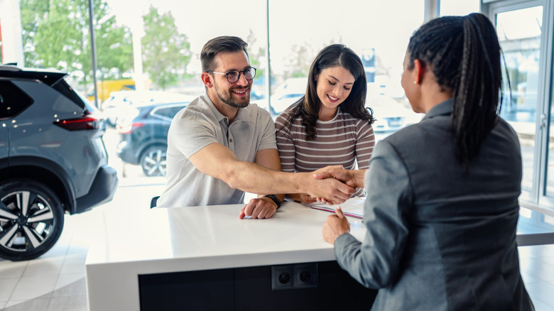 A happy couple shakes hands with a dealer representative over the desk at a large windowed dealership to confirm the sale.
