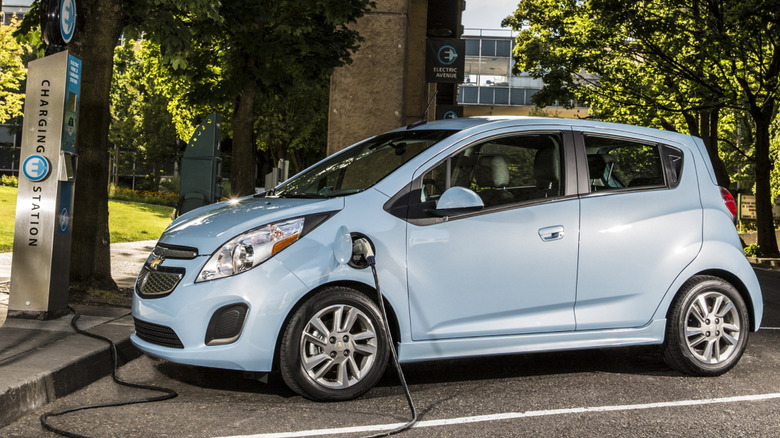 A side view of a light blue Chevrolet Spark EV parked and charging at a public charging station in front of trees