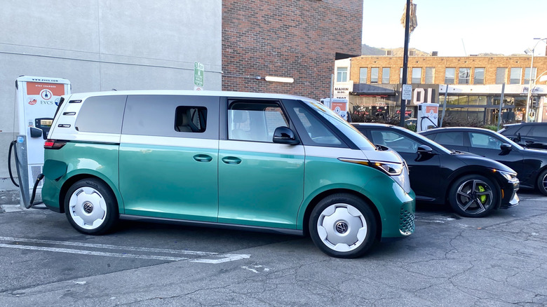 A side view of a green and white VW ID Buzz parked and charging at a public charger next to a black Kia EV6 GT