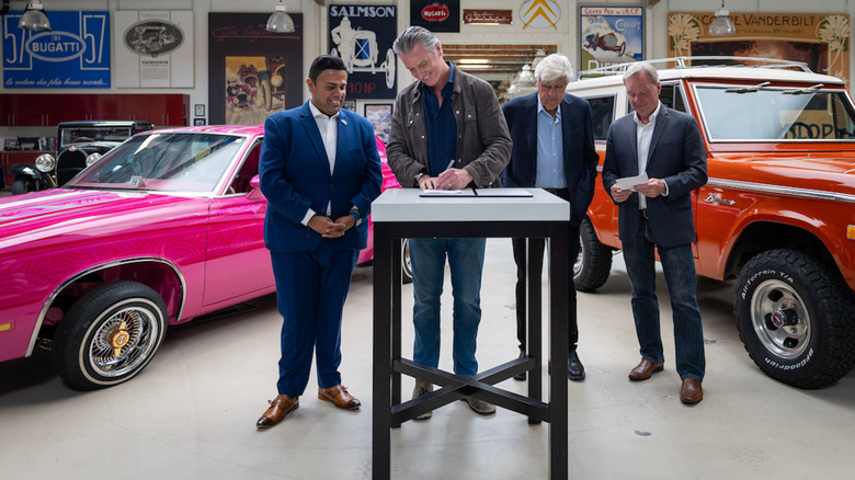 A photo of Gov. Newsom standing between a pink lowrider and an orange classic Bronco in Leno's garage signing the bills with three men around him