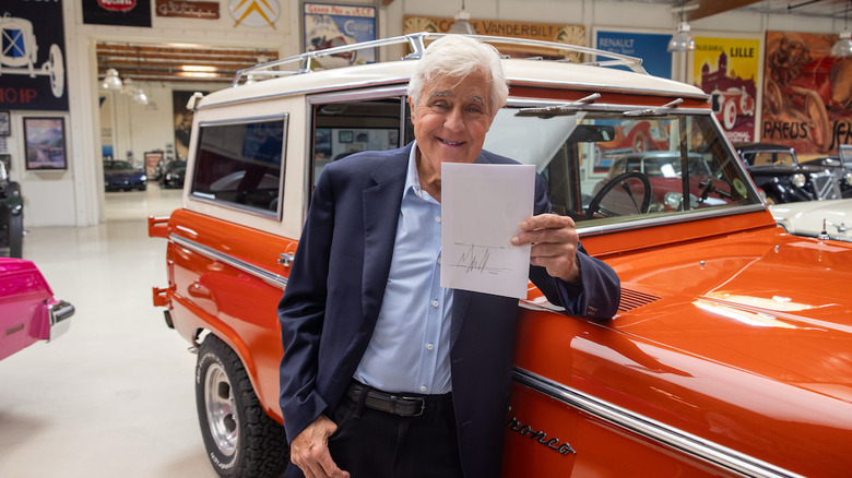 Jay Leno standing with his elbow on the orange Bronco in his garage holding the signed bill