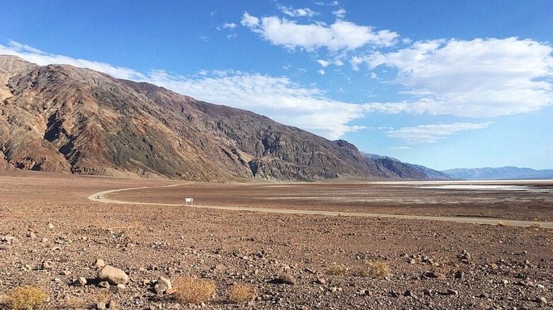 A curved section of Badwater Road in California cuts through the desert in front of a mountain, with a cloudy blue sky in the background