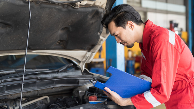 A car being inspected by a technician wearing a red jumpsuit and carrying a blue clipboard