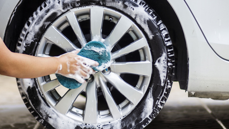 A car owner using a sponge and soapy water to clean the wheel and tire of a sedan