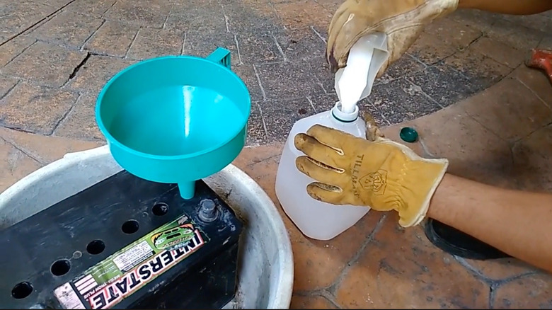 A person with gloves pours baking soda into a jug of water next to a battery with a funnel in it