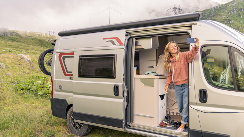Traveling in nature, a solo woman traveler takes pictures with her phone while standing in the sliding door of her RV