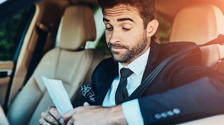 A man in a black suit site behind the wheel looking unhappily at his speeding ticket.