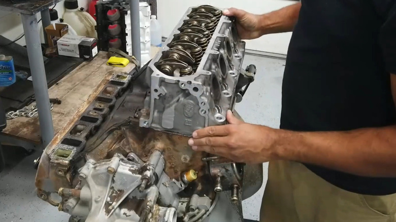 A man wearing a black t-shirt and khaki pants places an aluminum GM Chevrolet LT V8 engine head on a Ford Windsor 351 small block V8 block