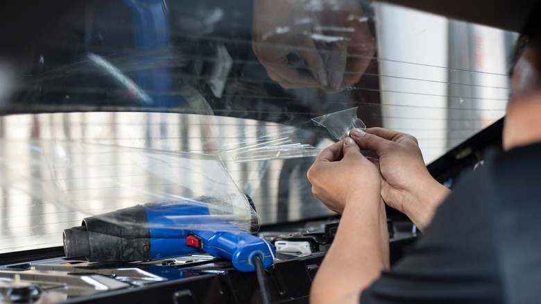 A person removing rear windshield tint using a heat gun while carefully peeling it off.