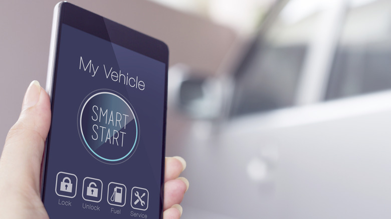 A cell phone in a woman's hand shows a remote car start feature, and a blurry gray car sits in the background