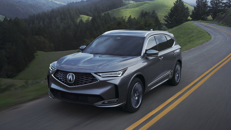 Front 3/4 view of a silver 2025 Acura MDX with the Advance Package navigating a sharp curve on a scenic mountain road.