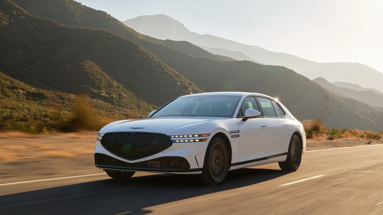 Front 3/4 view of a white Genesis G90 luxury sedan cruising along a mountainous highway during the golden hour.