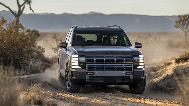 Front 3/4 view of a bronze 2026 Hyundai Palisade ICE XRT Pro navigating a dusty desert trail
