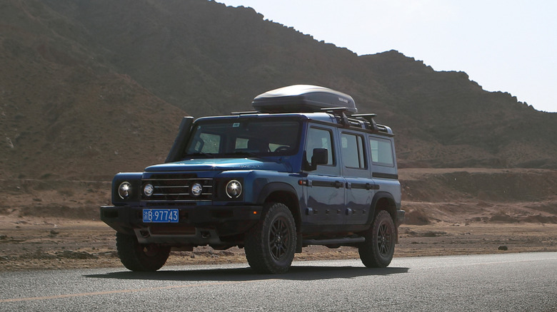 Front 3/4 view of a blue Ineos Grenadier equipped with a roof cargo box driving through a desert landscape.
