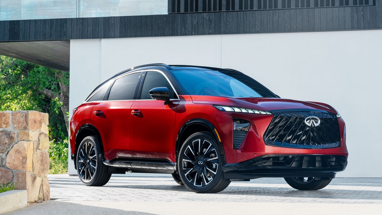 Front 3/4 view of a 2027 Infiniti QX65 in "Sunfire Red" paint parked on a light-colored stone driveway.