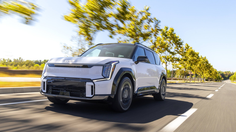 Front 3/4 view of a white 2026 Kia EV9 electric SUV driving fast on a sun-drenched road.