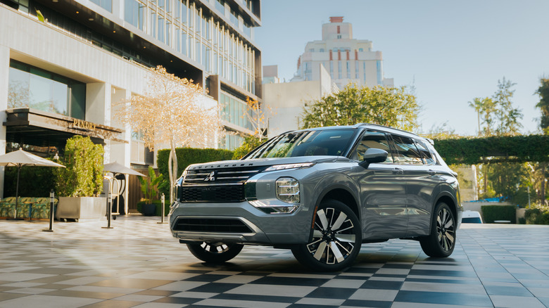 Front 3/4 view of a silver Mitsubishi Outlander parked on a checkered stone plaza in a modern urban setting.