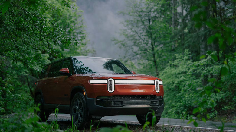 Front 3/4 view of a red Rivian R1S electric SUV parked on the side of a damp road, framed by a dense, lush green forest.