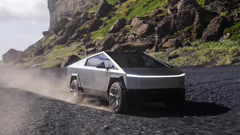 Front 3/4 view of a stainless-steel Tesla Cybertruck kicking up dust as it traverses a dark, volcanic beach.