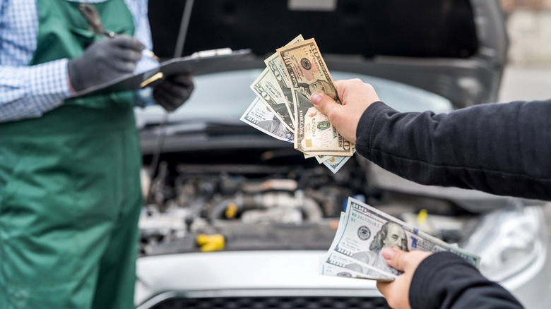 Two hands giving money to an auto tech with a car with an open hood in the background