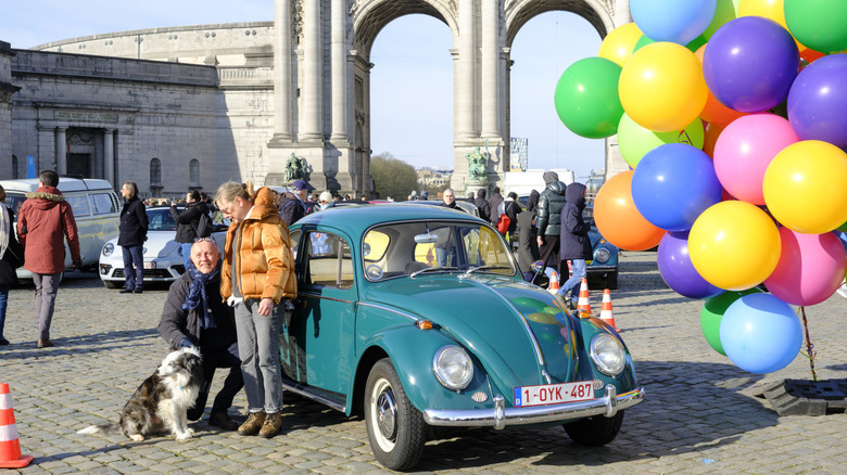 A teal VW Bug parked on cobblestones next to a bunch of colorful balloons. A nice older couple stand in front of the car with their shepherd dog