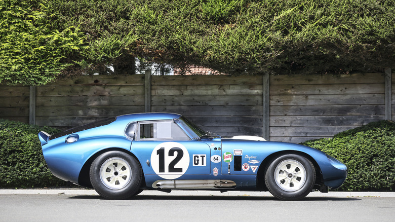 The beautiful blue 1964 Shelby Cobra Daytona Coupe wears multiple stickers from its former races and the number 12 in a white circle, pictured against a wooden fence surrounded in greenery.