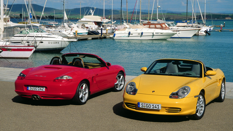 A red and yellow 986 Porsche Boxster S parked on a sandy area with boats nearby.