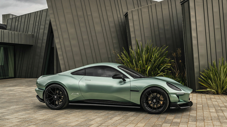A side view of a light green Project V parked on bricks in front of a black structure and plants
