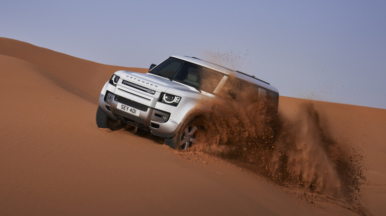 A white Land Rover Defender throws sand while tearing up a dune.