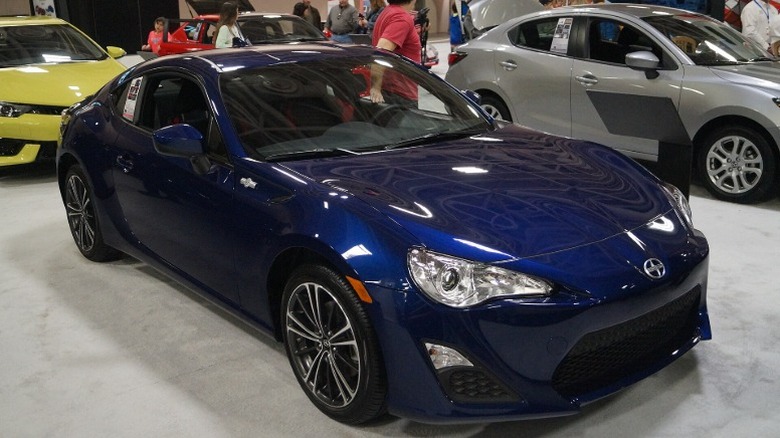A blue 2016 Scion FR-S parked at an auto show with people and other cars in the background