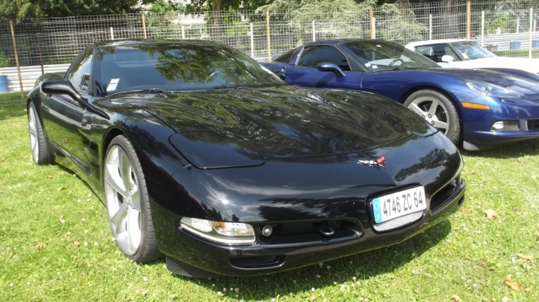 A black C5 Corvetter parked in a grass lot in front of an iron mesh fence.