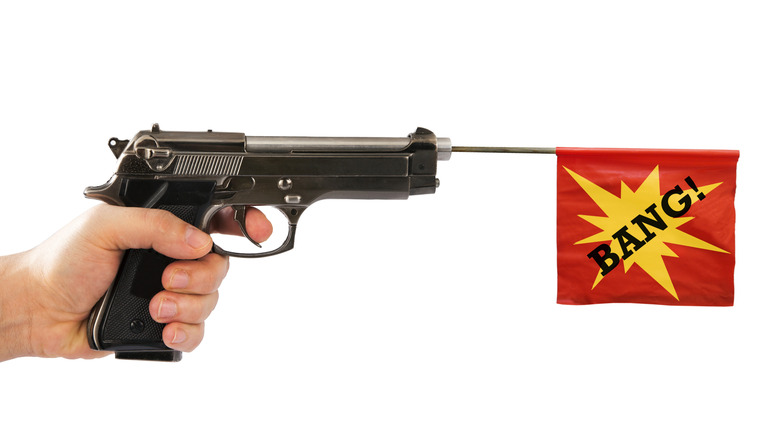 A man's hand holding a Beretta 92 or M9 pistol with a Bang! flag coming out of the barrel