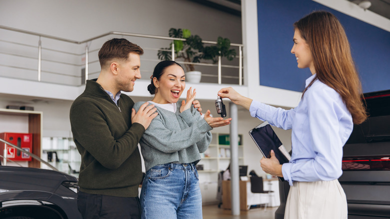 Excited couple receiving new car keys from salesperson at automobile dealership showroom, celebrating purchase