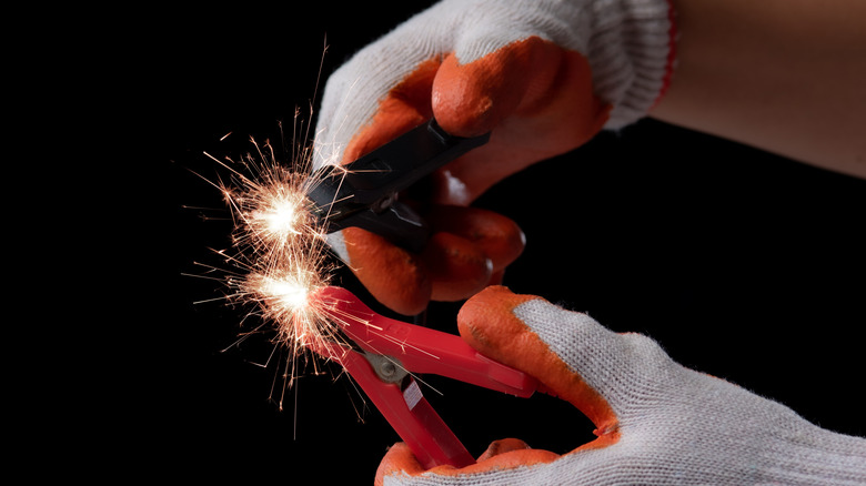 A person wearing protective gloves touching both negative and positive jumper cable clamps together.