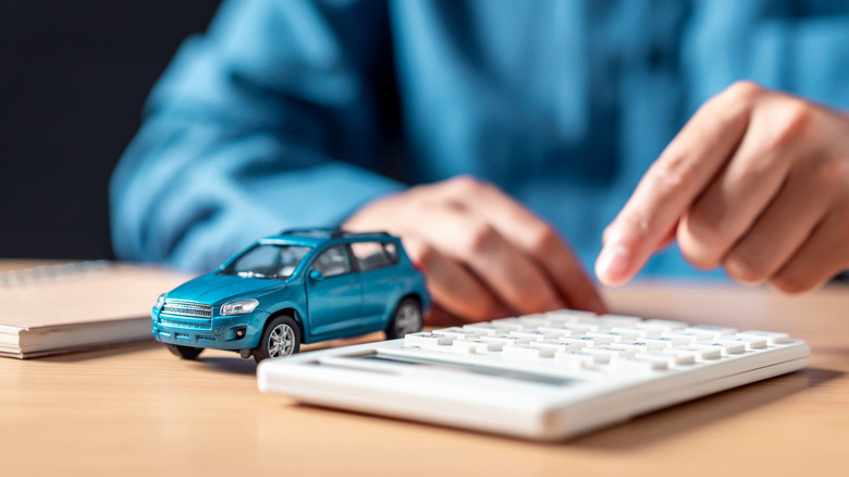 A blue toy car sitting next to a calculator with a person's hand pointing at it.