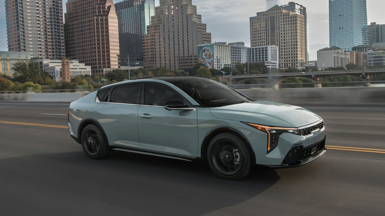 A Kia K4 driving on the highway in front of a city skyline.