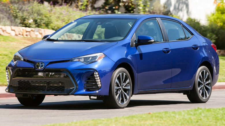 Toyota Corolla driving on a paved road with trees and buildings in the background