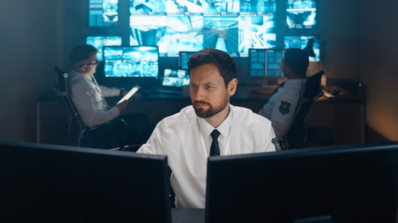 A uniformed man sitting in front of computer screens with other officers and large screens in the background