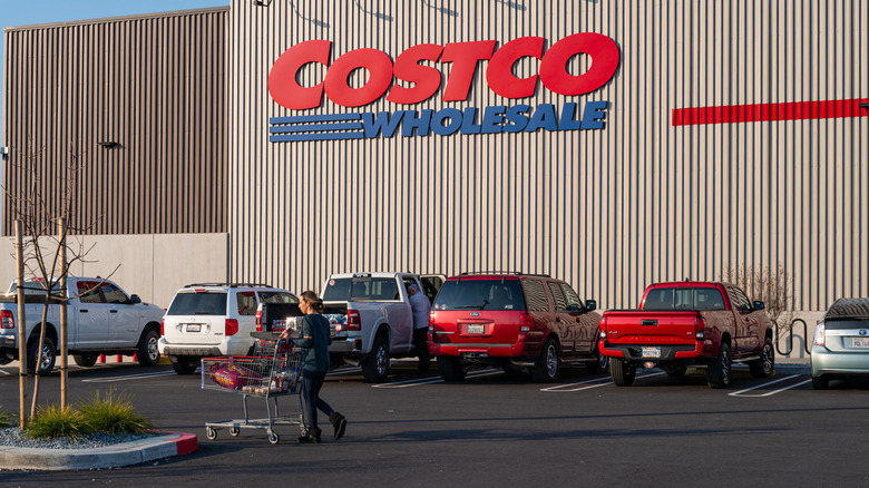 A shopper returns to their vehicle with a cart full of products, with a row of parked vehicles and the Costco logo in the background.