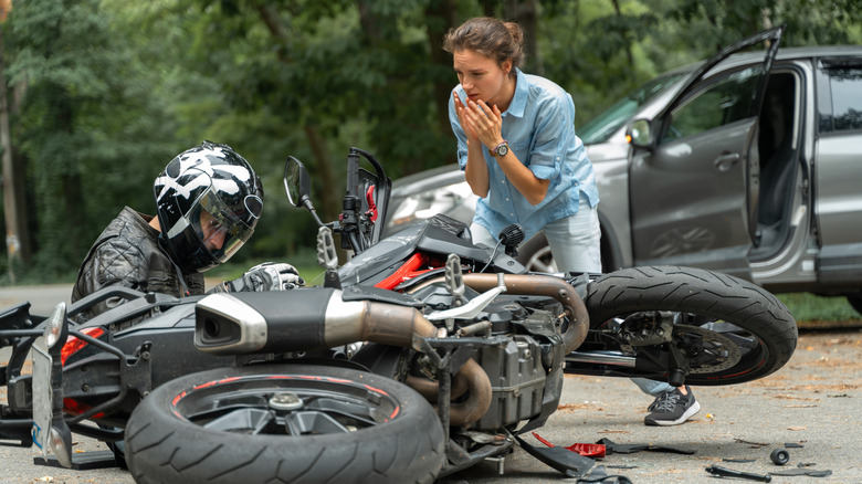 A scene of an accident between a car and a motorcycle, with the motorcyclist sitting next to his wrecked bike as the driver comes over to check on him.