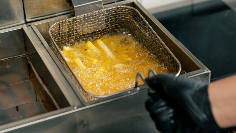 A black gloved hand holds a basket full of french fries in a deep frier full of vegetable oil
