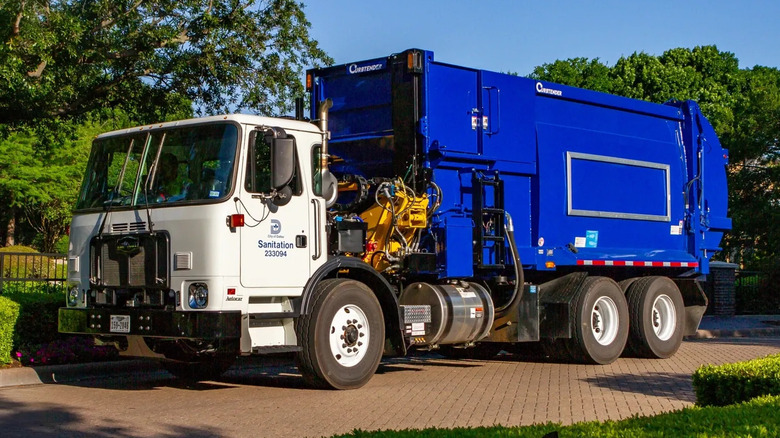 Front three quarters shot of a Dallas Sanitation Department trash truck with a white cab and blue container on the back parked on brick in front of trees and a blue sky