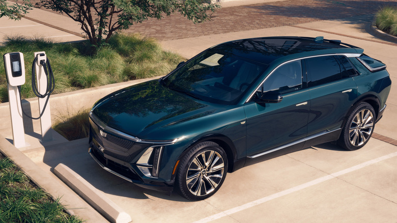 A front three quarters overhead shot of a dark green Lyriq parked on concrete at an EV charger, though it's not plugged in