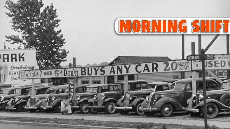 Automobiles on display at a second-hand car lot on Queens Boulevard, New York, 1930s. A man is examining the front of a car, below a sign that reads 'Why Walk? When $5 Down Buys Any Car, 20 Months To Pay'.