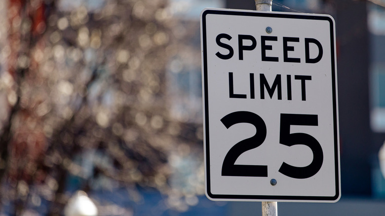 A close-up of a standard white regulatory speed limit sign showing 25 MPH in bold black text, mounted on a metal pole along a suburban city street with blurred residential buildings and trees in the background.