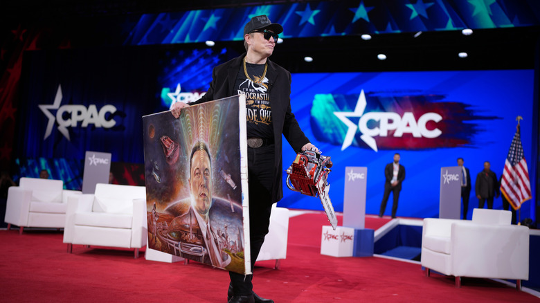Elon Musk at CPAC in 2024, holding a painting of himself in one hand and a chainsaw in the other