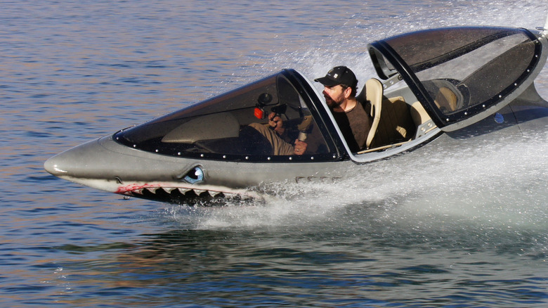 A man sits in the open cockpit of a shark model, the hand controls are easily viewable, as the shark skims across the water.
